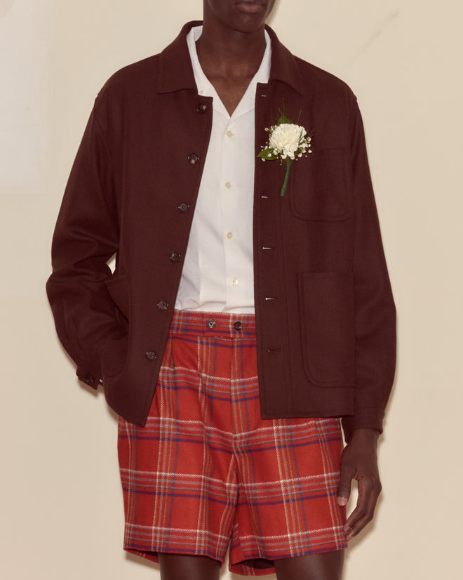 A person wears a Bramble Franck Jacket in dark brown wool twill, paired with a white shirt, red plaid shorts, and a white flower pinned to the jacket.