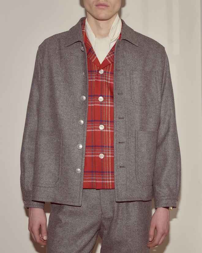 Someone stands against a plain background, wearing the Dovecote Franck Jacket in heathered grey wool, layered over a red plaid vest and a white shirt.