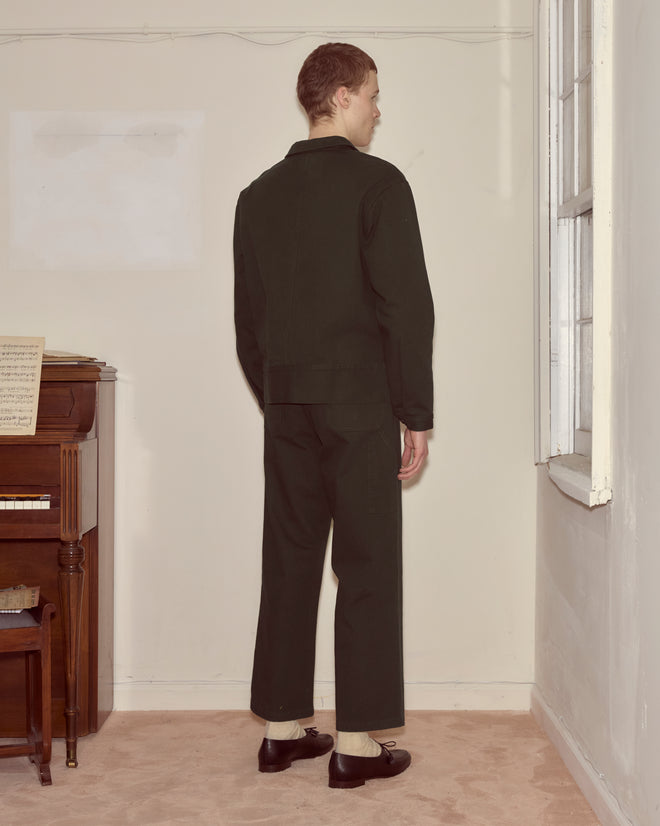 A person wearing the Twill Knolly Brook Side Tab Jacket in Dark Green stands by a window near a piano with sheet music, facing a corner.