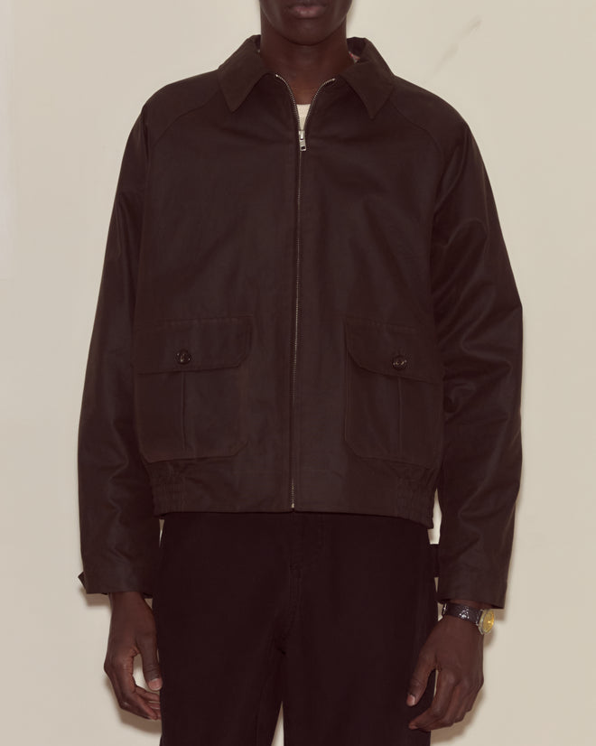 A person wearing the Waxed Coach’s Jacket - Brown, featuring front pockets, stands against a plain background. This jacket combines timeless style and practical functionality.