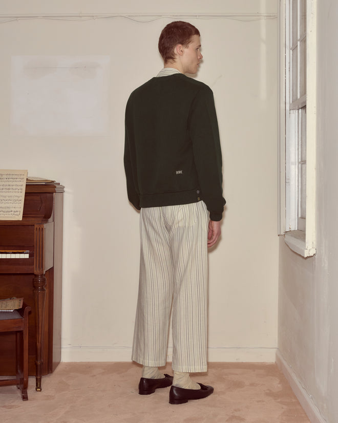 A person stands in the corner of a room, wearing a dark green Double-Breasted Cardigan and striped pants near a piano.
