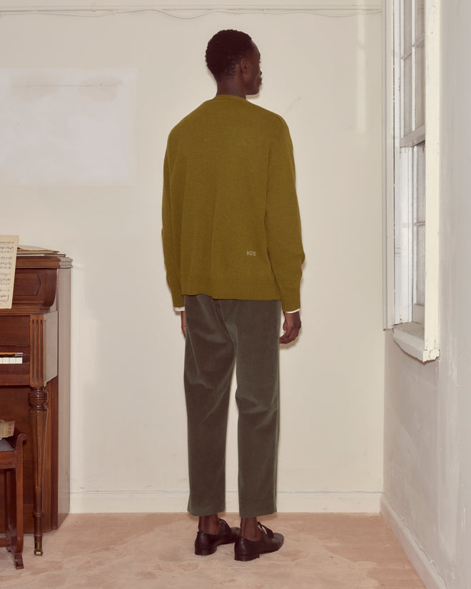 A person wearing the Orchard Cardigan - Green stands indoors facing a wall near a piano and a window.