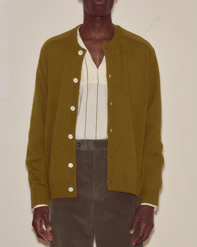 Person wearing the Orchard Cardigan - Green over a white shirt and olive corduroy pants, standing against a beige wall.