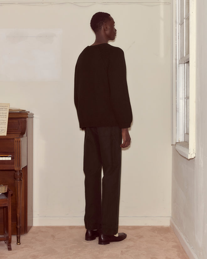 Dressed in a black Cashmere Crewneck with an oversized silhouette, a person stands facing a cream wall next to a piano and window.
