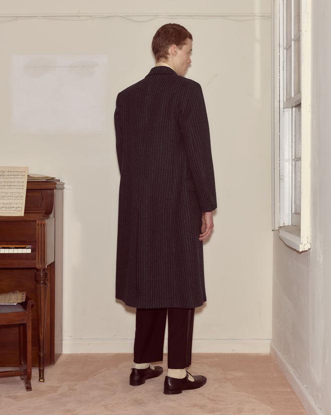 A person wearing the Waldorf Pinstripe Coat stands facing a corner near a piano and window in a sparsely furnished room.