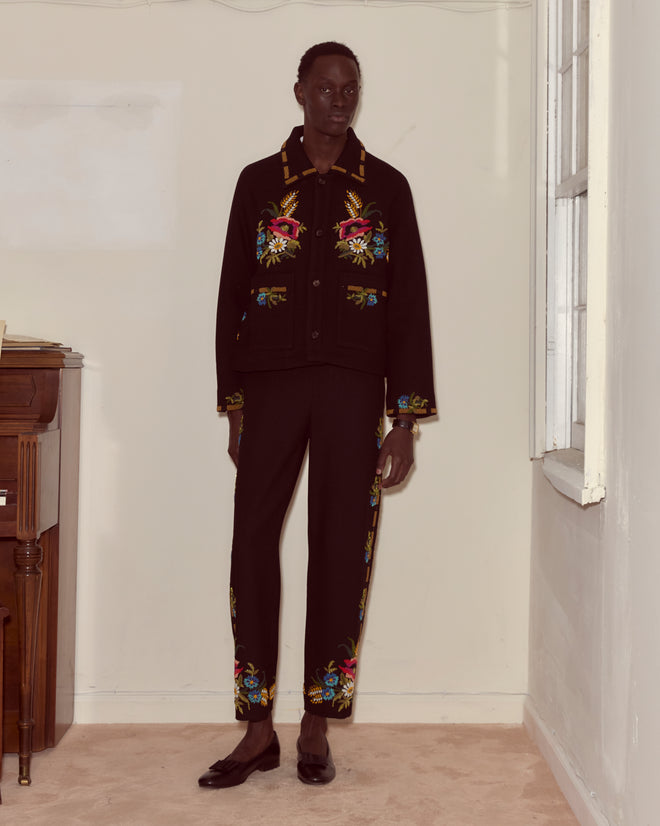 A person wearing the Beaded Aquitaine Jacket, featuring vibrant floral embroidery, stands indoors by a window.