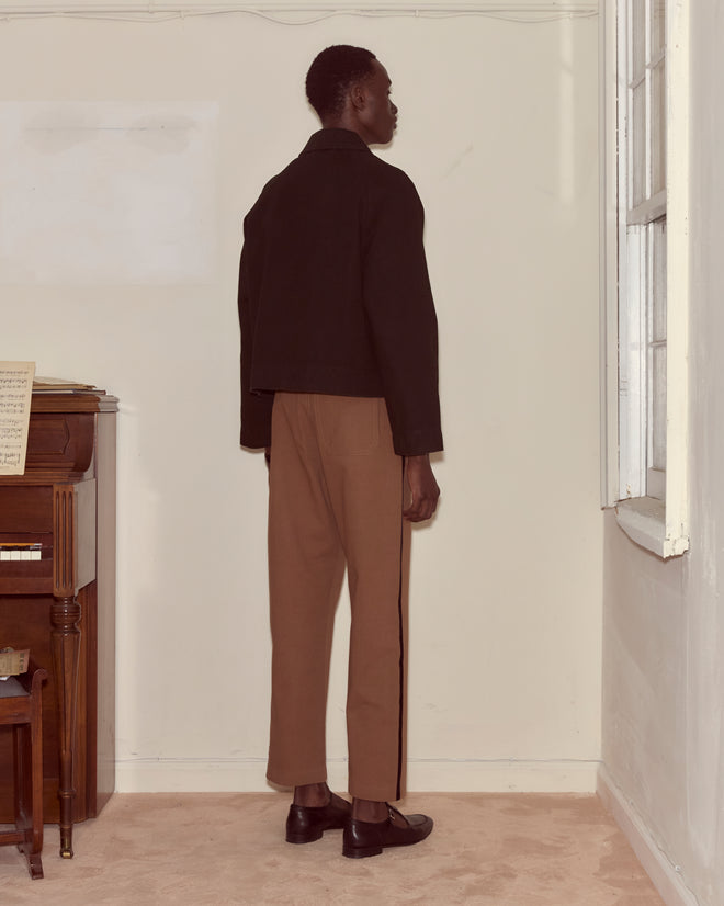A person stands facing a corner in a room, wearing a black Embroidered Denim Jacket with a boxy fit, brown pants, and black shoes.
