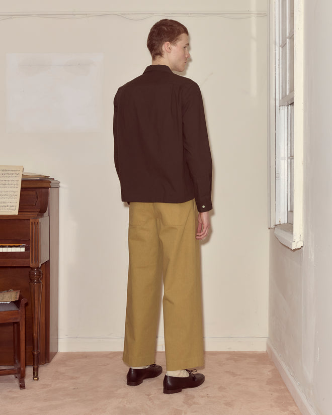 A person wearing a brown Voile Long Sleeve Shirt stands indoors by a piano and window, facing away.