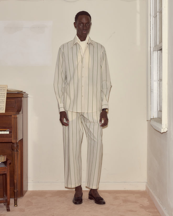 A man stands indoors in a cream pinstripe suit over the Celery Stripe Long Sleeve, paired with black shoes beside a piano and window.