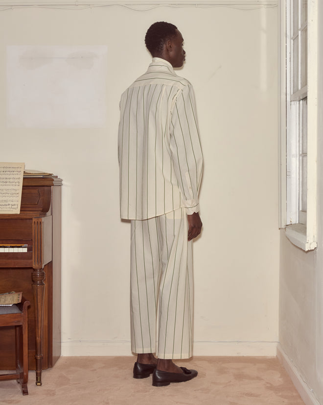 Dressed in the Celery Stripe Long Sleeve, a person stands by a piano, facing the wall in a softly lit room, evoking the style of 1930s shirting with its cream pinstripes.