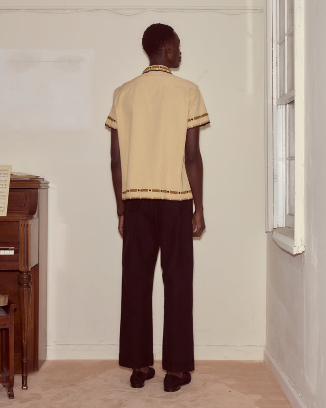 A person stands in a corner, wearing the Beaded Ryegrass Short Sleeve Shirt with handmade beadwork and black pants.