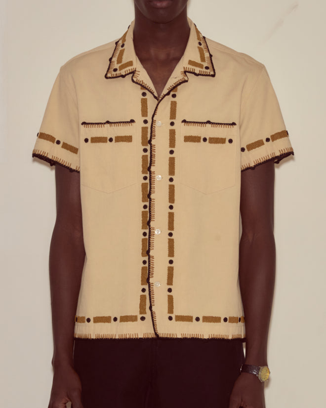A person models the Beaded Ryegrass Short Sleeve Shirt, featuring beige fabric and brown and black stitched patterns, while standing in front of a plain wall.