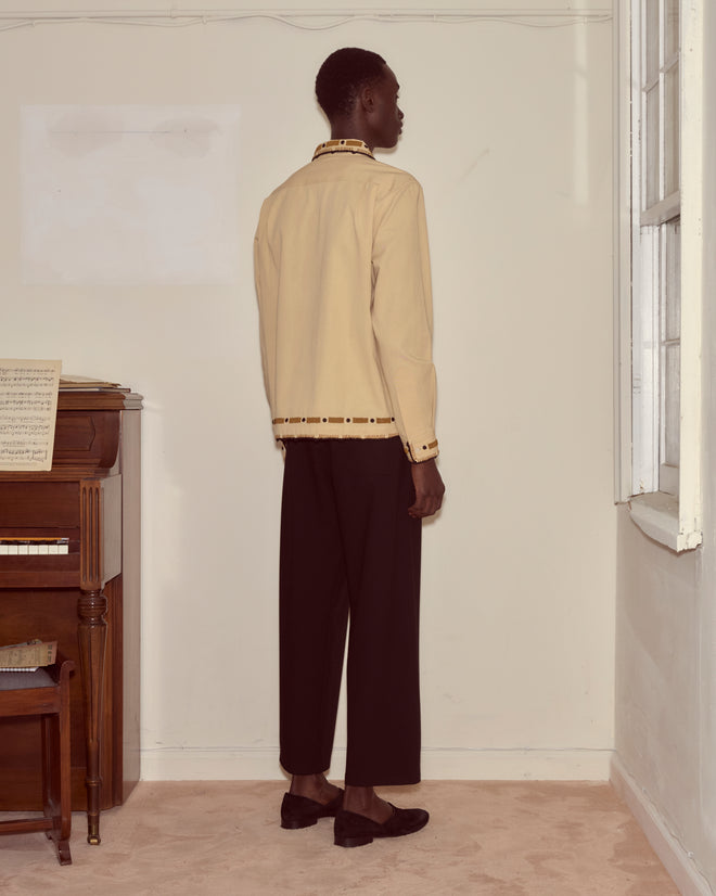 A person stands by a piano facing a blank wall, wearing a beige jacket, black pants, and the Beaded Ryegrass Long Sleeve Shirt with intricate handmade beadwork.