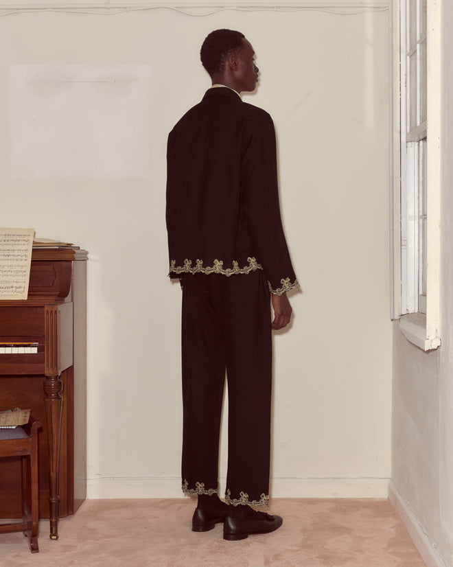 A man wearing the Laurelist Overshirt - Black with gold wreath-and-garland trim stands facing a wall near a piano and window.