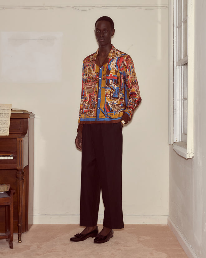Wearing a Manhattan Island Long Sleeve Shirt and black pants, a person stands indoors by a piano and window.