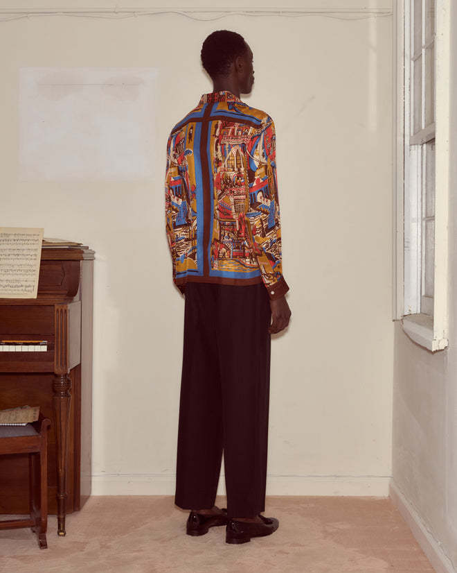 A man in a Manhattan Island Long Sleeve Shirt and black pants stands facing a cream wall near a window and piano.