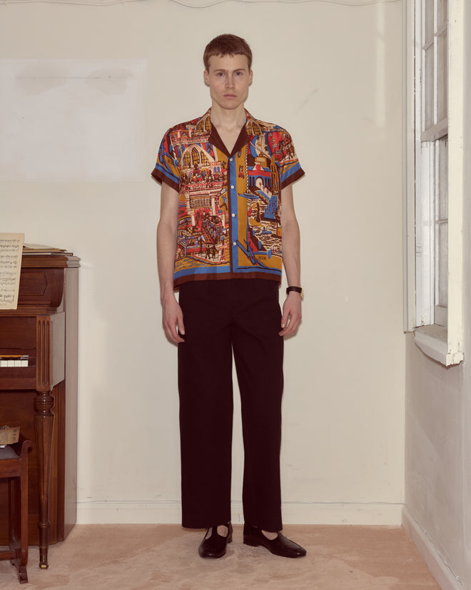 A person stands indoors next to a piano wearing the Manhattan Island Short Sleeve Shirt, black pants, and black shoes.