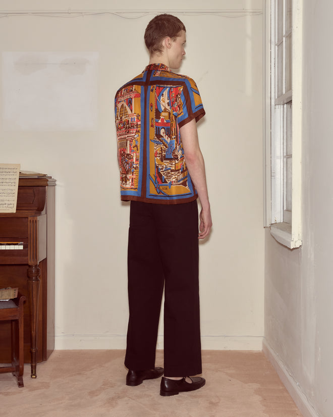 Wearing the Manhattan Island Short Sleeve Shirt and black pants, a person stands facing a corner near a window and upright piano.
