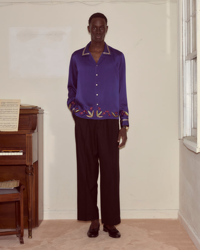 Indoors, a person stands beside a piano, wearing the Dream Souvenir Long Sleeve—a blue textured silk shirt with embroidery—paired with black pants.