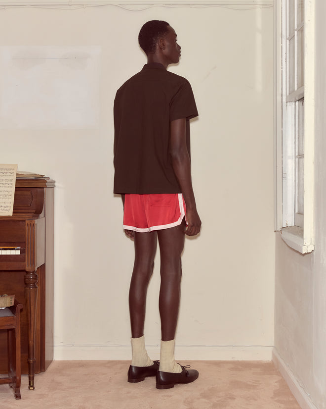 A person in red shorts, the brown Voile Short Sleeve Shirt with a boxy silhouette, and socks with shoes stands by a window, facing away in a room.