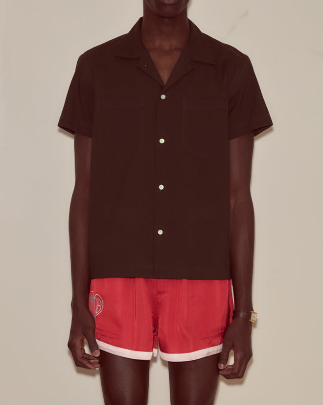 A person wearing a brown voile short sleeve shirt with a boxy silhouette and red athletic shorts stands against a plain background.