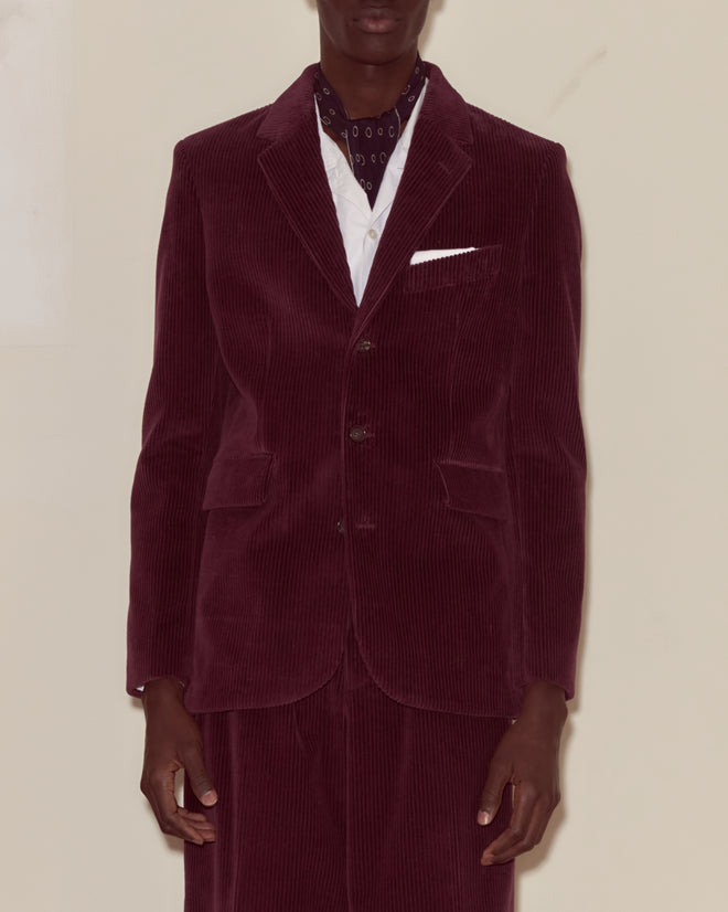 A person wears a dark purple Corduroy Suit Jacket with a white shirt and patterned scarf, channeling 1950s men's fashion against a plain background.