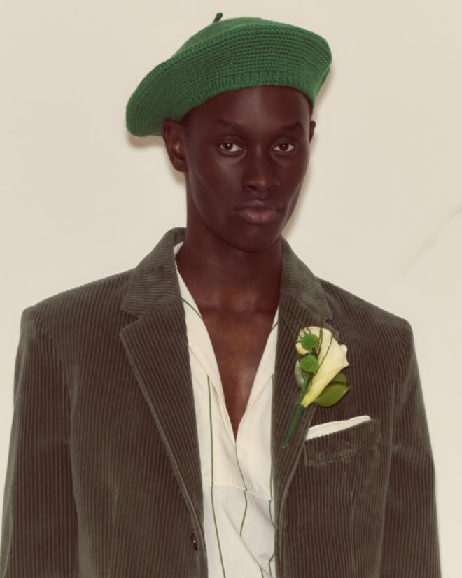 A person wearing a green beret and the Corduroy Suit Jacket - Green, styled in 1950s fashion with a white rose boutonniere, looks at the camera.