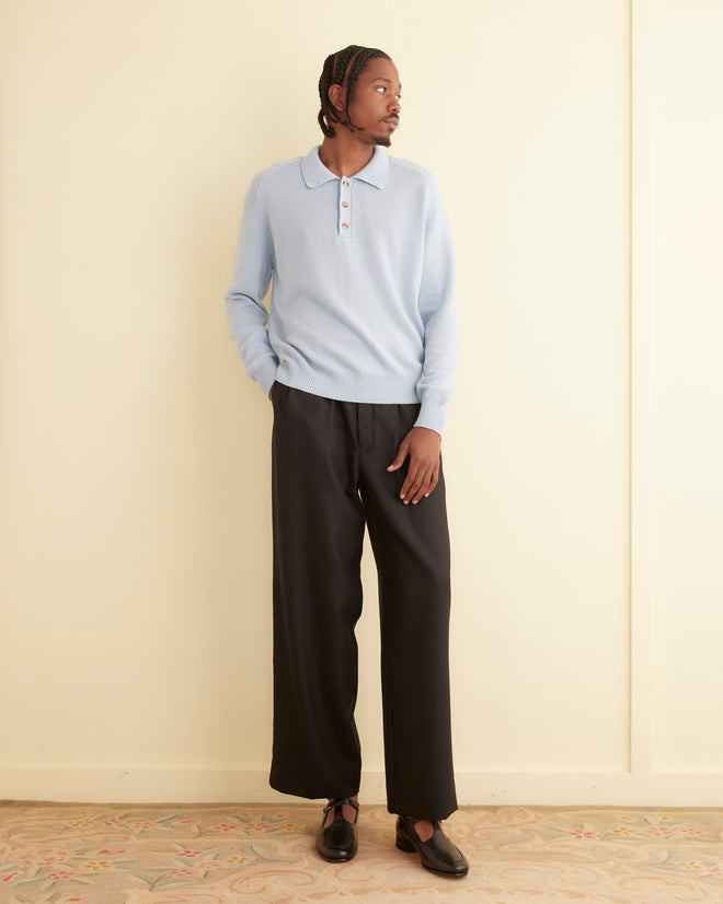 A person wearing a light blue Cashmere Polo and black pants stands with hands in pockets, looking to the side.