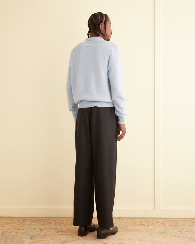 A person with braided hair, wearing a light blue Cashmere Polo and dark pants, stands facing away in a softly lit room.