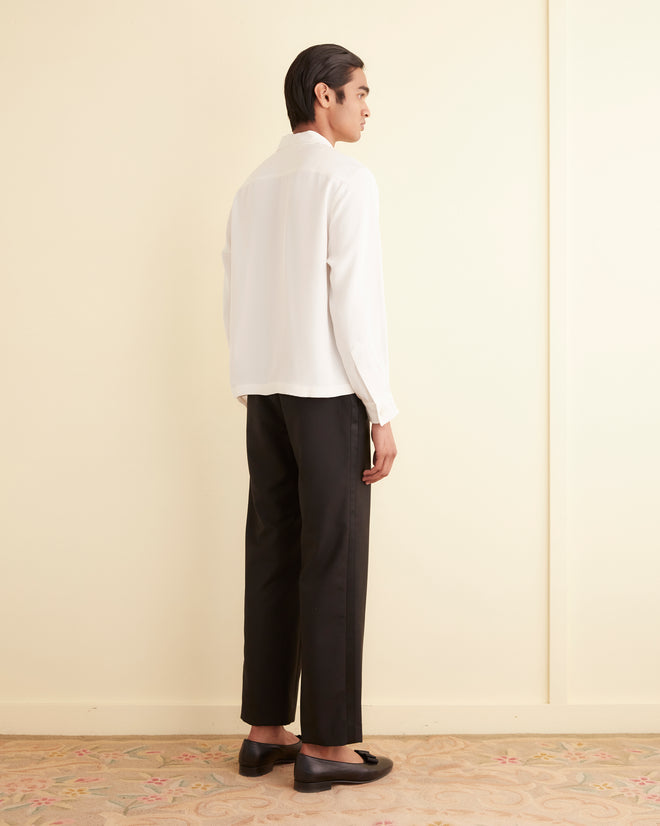 Person with short dark hair stands sideways indoors, wearing the Trillium Long Sleeve Shirt in white, 1940s-inspired silk crepe with trompe l'oeil closures, paired with black pants and black shoes.