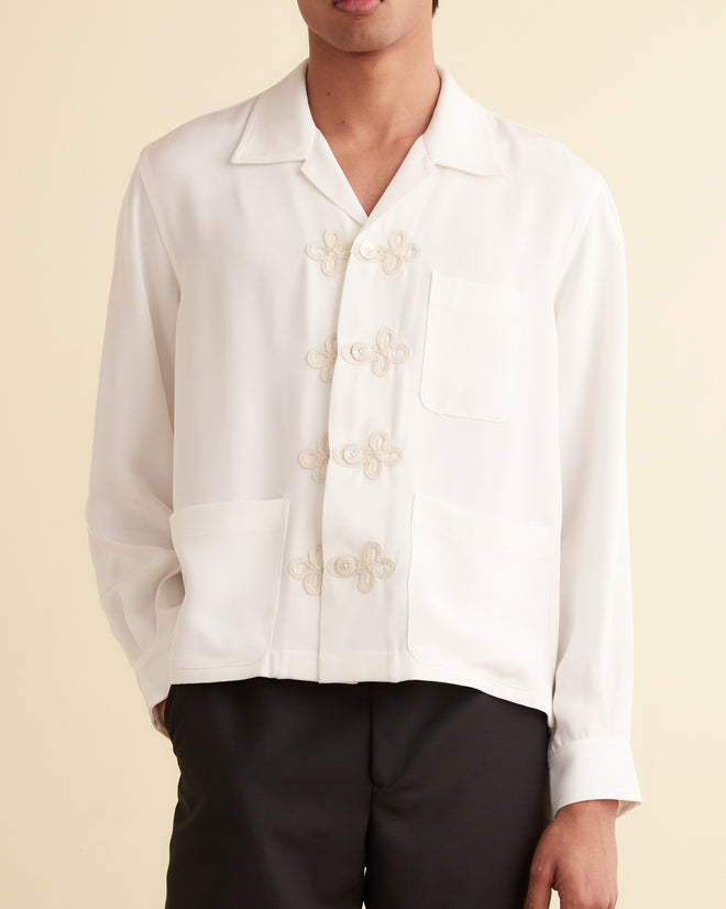 Man wearing the Trillium Long Sleeve Shirt in white, featuring 1940s-inspired details and decorative knot buttons, paired with black pants against a beige background.