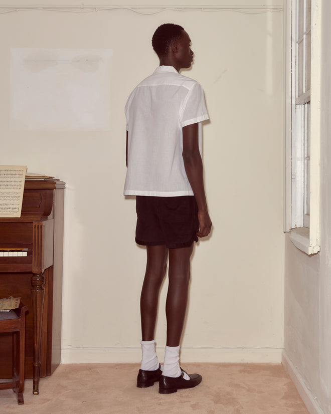 Wearing the Voile Short Sleeve Shirt - White and black shorts, a person stands by a window and piano with sheet music, facing a beige wall and showcasing the shirt's relaxed, boxy silhouette.