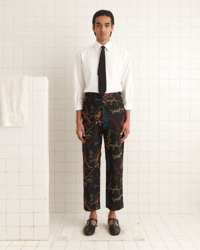 A person in a white shirt and tie wears the mid-rise Signature Scrawl Trousers, featuring colorful patterns and made from 100% cotton, on a tiled floor.
