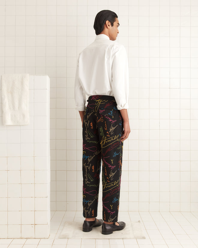 A person in a white shirt and Signature Scrawl Trousers stands in a tiled room with a 100% cotton towel on a rack.