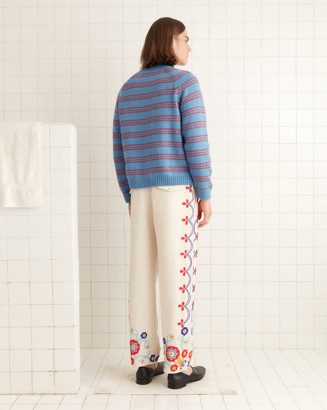 In a tiled room, a person stands facing away wearing a blue striped sweater and the Beaded Garden Party Trousers, crafted from 100% linen and featuring colorful floral embroidery.