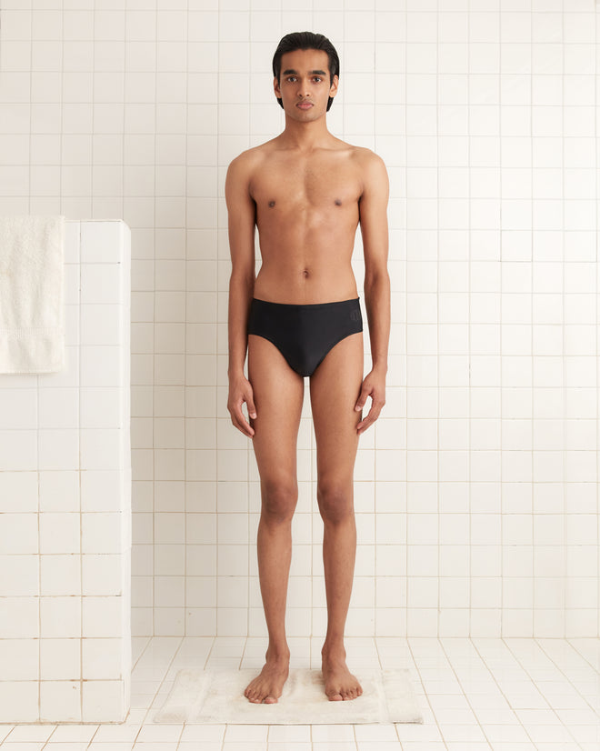 A person stands in a tiled room wearing Swimming Briefs - Black, featuring a visible Bode logo, facing forward with arms at their sides.