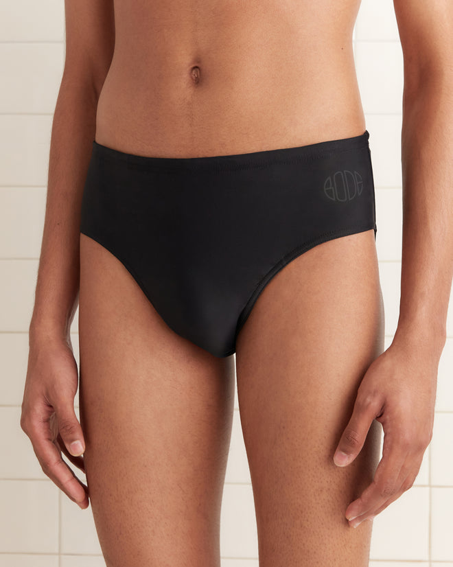 A person stands against a light tiled background wearing black Swimming Briefs - Black, a high-waisted men’s swimsuit with a subtle Bode logo.