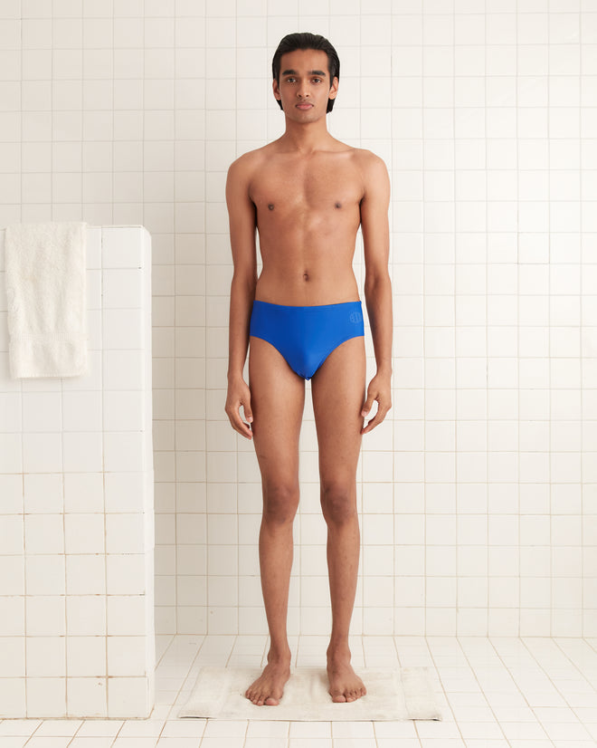 A person stands in a tiled bathroom, facing forward on a white bath mat, wearing Swimming Briefs - Blue made from recycled polyester.