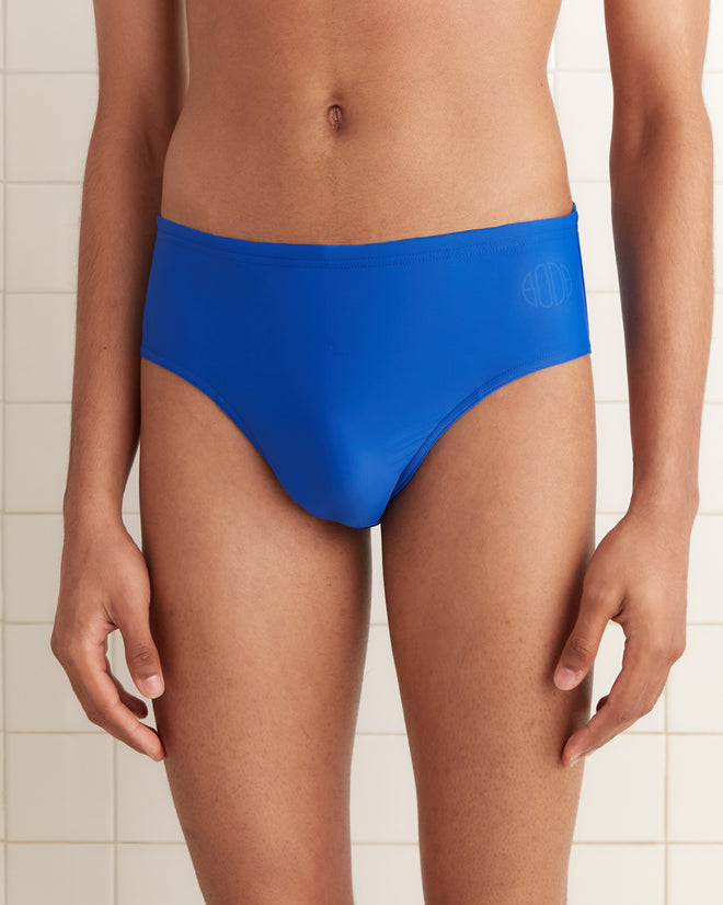 Someone in Swimming Briefs - Blue, crafted from recycled polyester, stands before a tiled wall, shown from shoulders to mid-thigh.