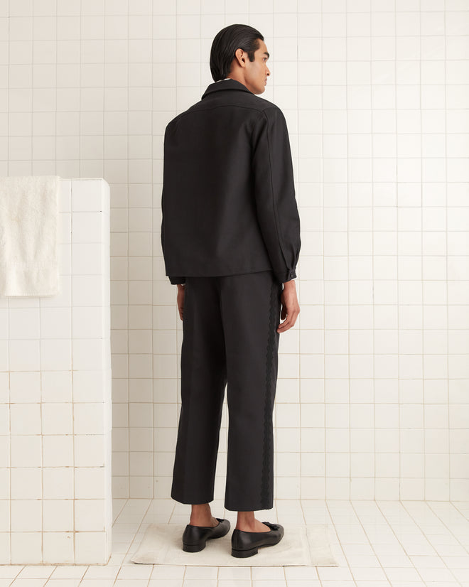 In the tiled room, a person in a black wool twill outfit wears the Seymour Franck Jacket, blending seamlessly as they face away.