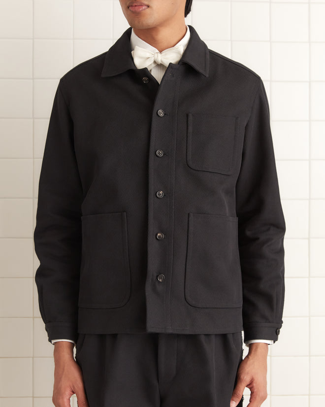 A person is standing against a tiled wall, wearing the Seymour Franck Jacket, a black wool twill men's workwear coat featuring large pockets.