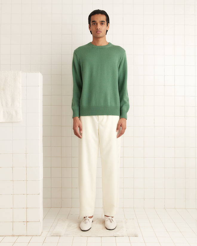 In a white-tiled room, someone wears the Juniper Sweater in green, a 1960s-inspired piece made from soft merino wool, paired with crisp white pants.