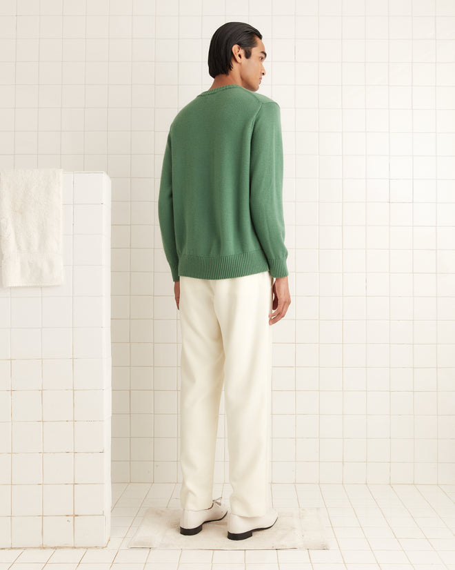 Someone wearing the Juniper Sweater - Green and white pants stands in a white tiled room, with the sweater adding a hint of retro charm to the scene.