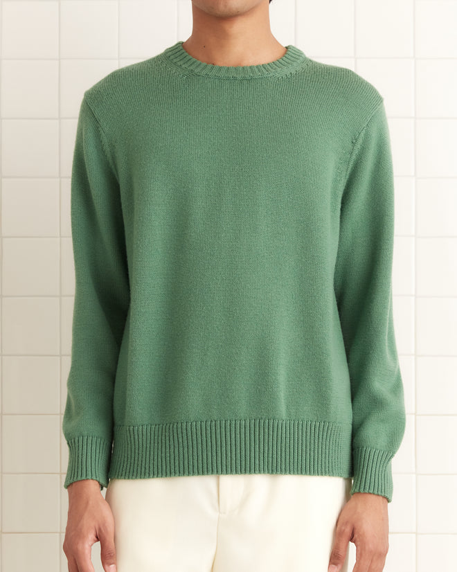 Someone wearing a Juniper Sweater in green and white pants stands elegantly against a tiled background.