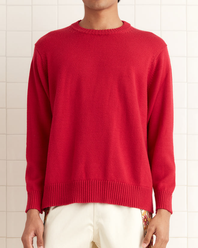 A person is wearing a Juniper Sweater in pink/red with white pants, posing against a tiled wall.