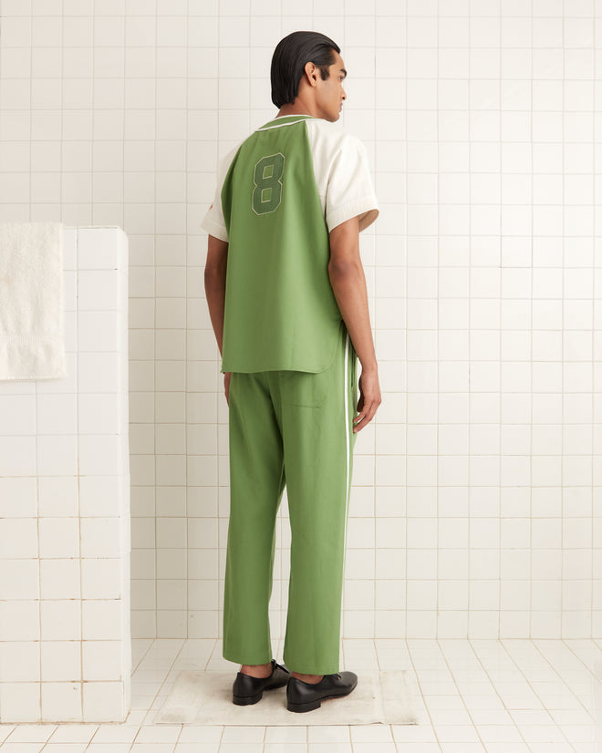 In a tiled room with white walls, someone wearing an Outfield Baseball Shirt in green and white, marked with the number 8 on the back, exudes a nostalgic 1940s vibe.