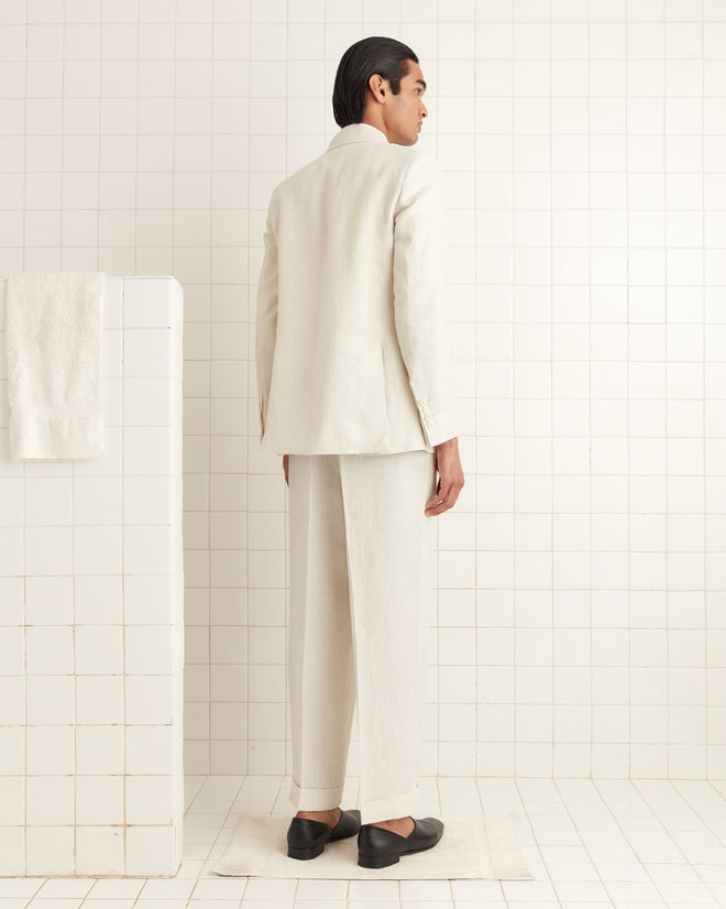 In a tiled room, a person in the Nelson Single-Breasted Suit Jacket, the beige color reminiscent of 1950s-60s style, faces away next to a half wall with a towel, evoking timeless elegance and mystery.