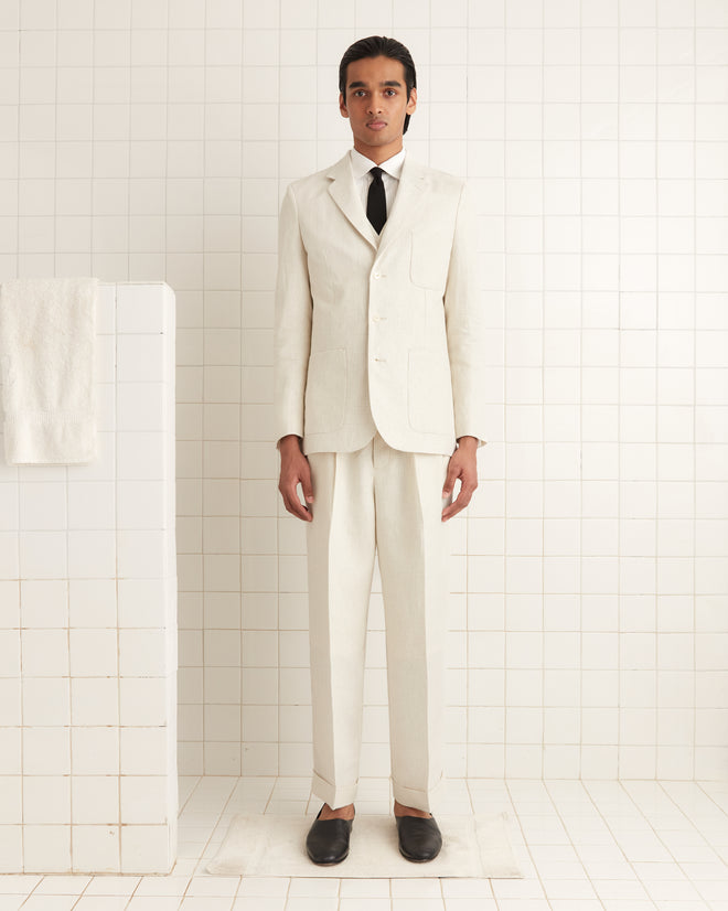 In a tiled room, someone elegantly stands in a 1950s-1960s outfit wearing the Nelson Single-Breasted Suit Jacket with an ivory taupe graph check pattern and a black tie, beside a towel on a rack.