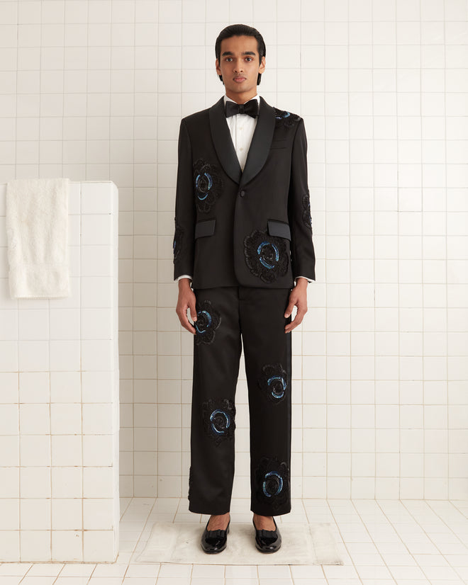 Dressed in the Poppy Shawl Collar Suit Jacket, a person exudes timeless sophistication in a 1940s-inspired, embellished black tuxedo with intricate beadwork, standing elegantly in a tiled room with a crisp white shirt and classic bow tie.