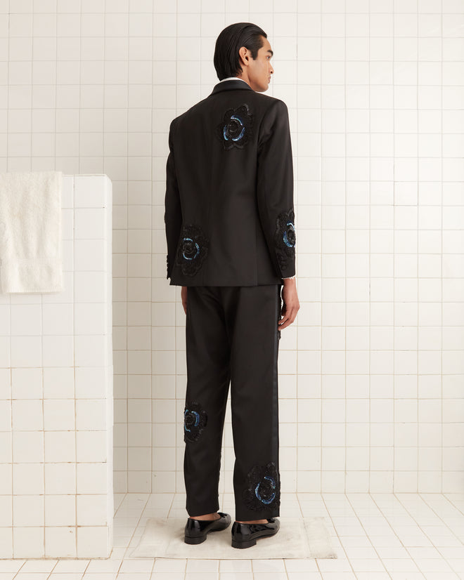 In a white-tiled room, a person wearing the Poppy Shawl Collar Suit Jacket, featuring intricate 1940s-inspired patterns, stands facing away.